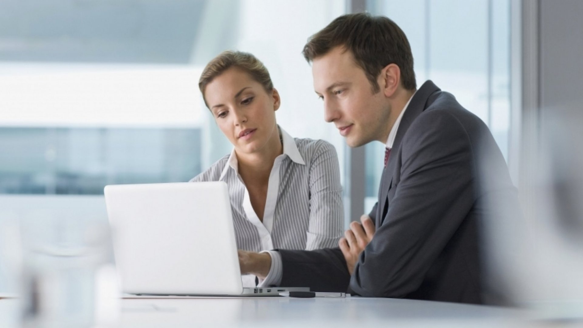 A man wearing a grey suit sitting beside a woman wearing a shirt as both of them stare at a laptop