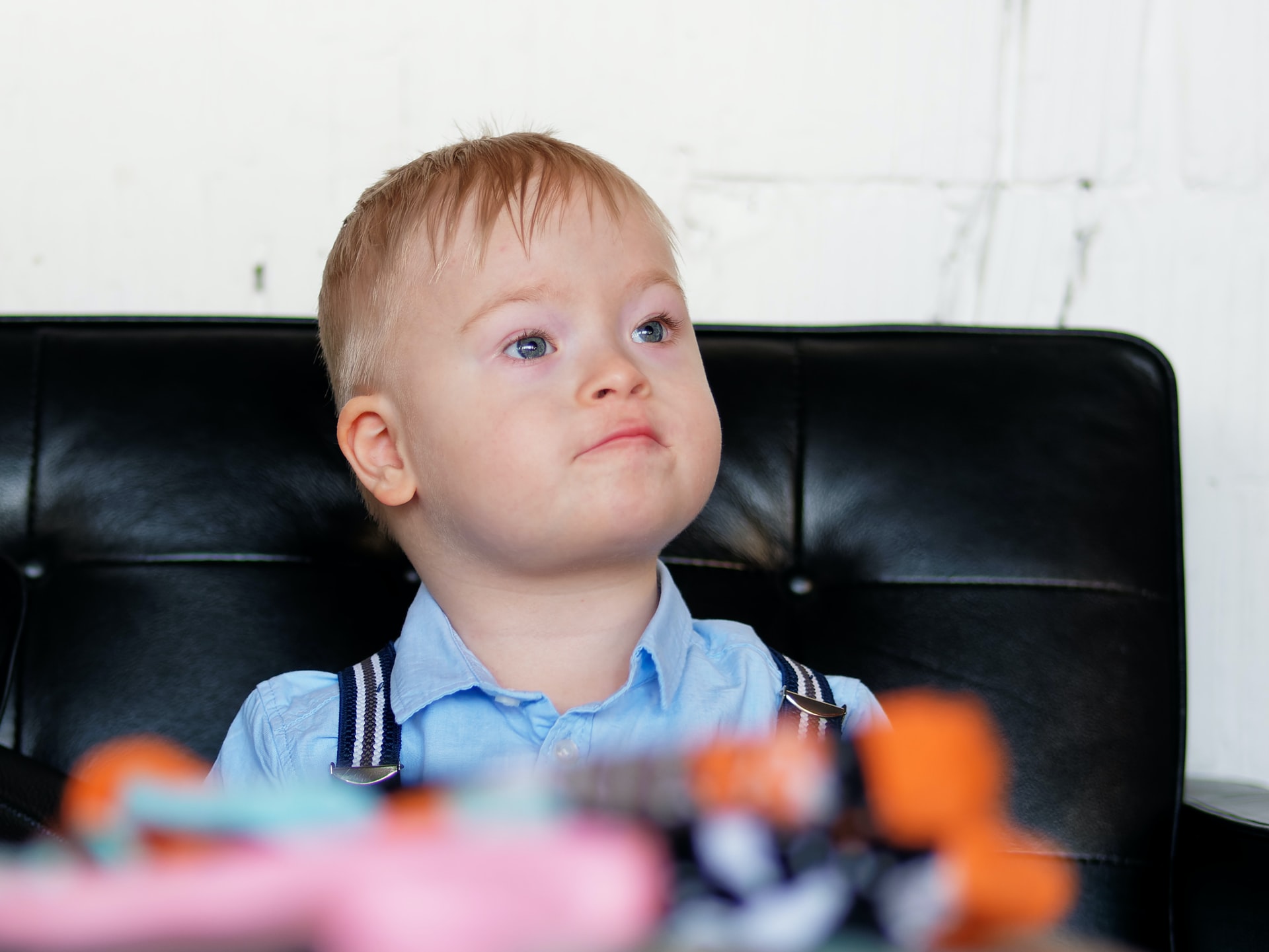 A baby in a blue shirt with a closed mouth is watching attentively while sitting on a black couch
