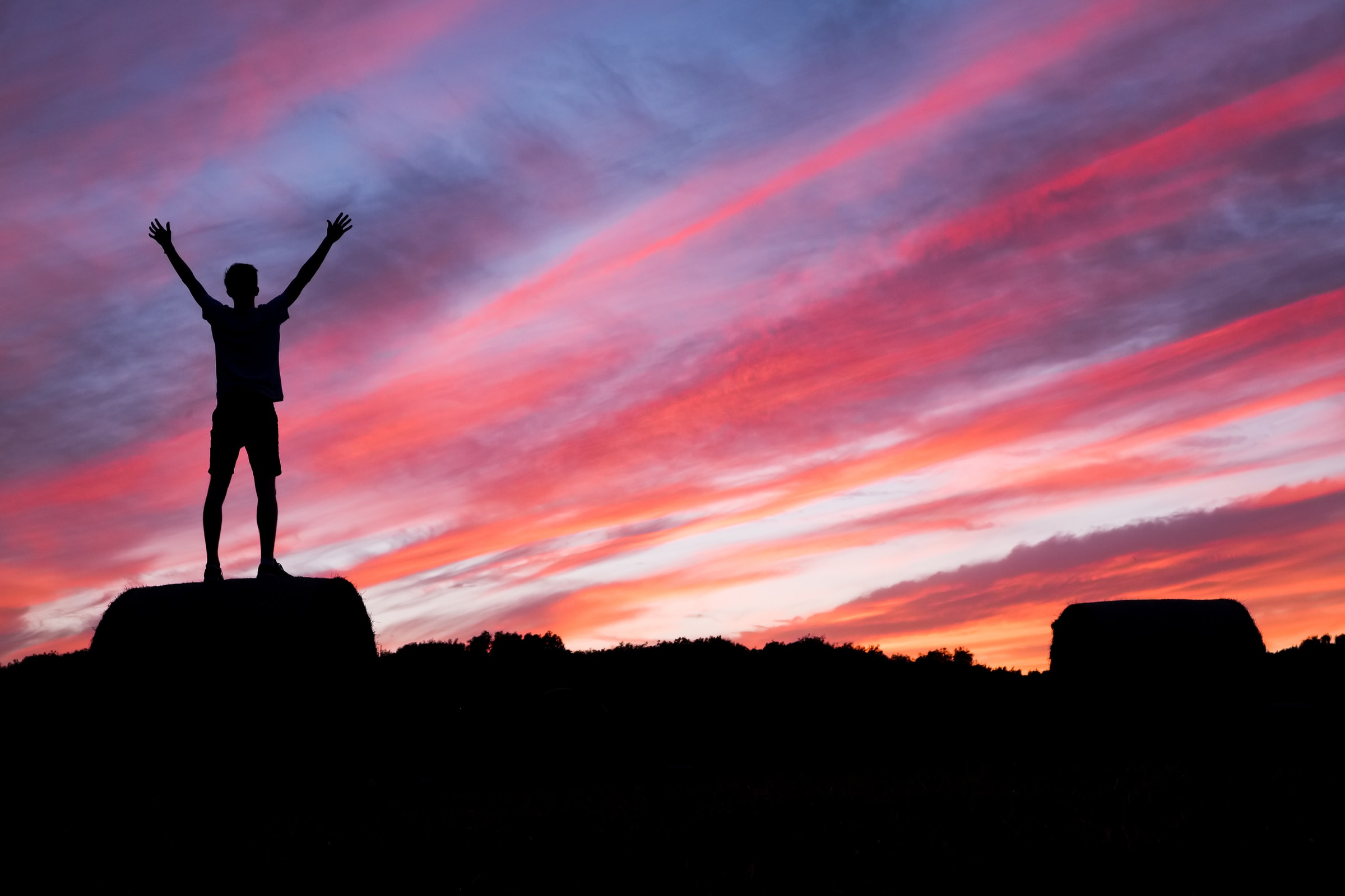 A man standing on the top of a rock at night with his two hands raised to a colorful night sky